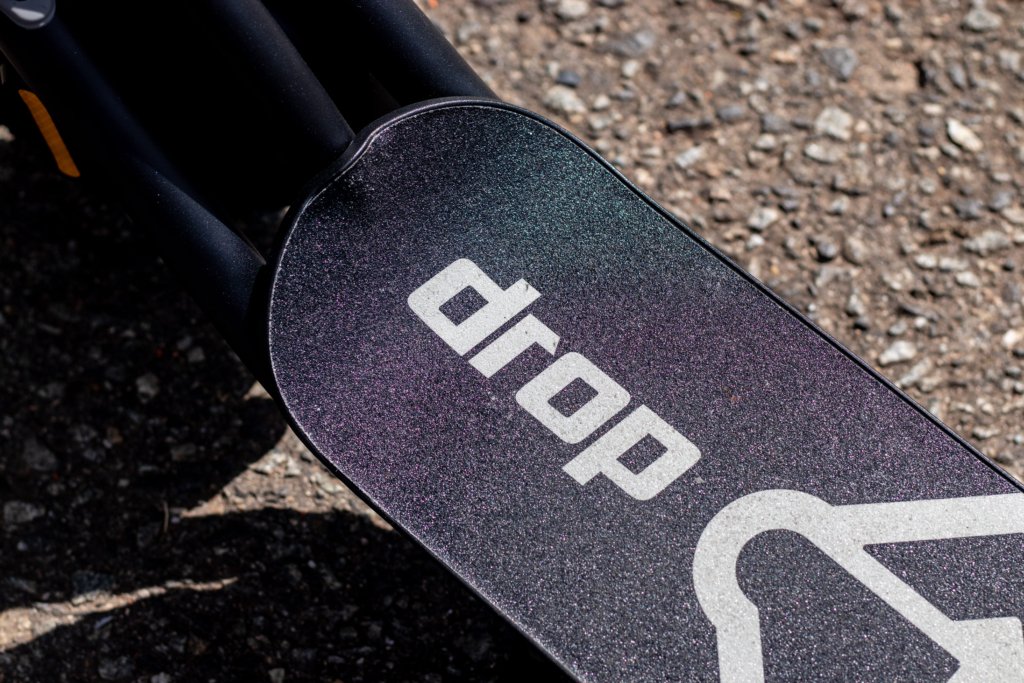 Patinete Elétrico para Adultos: tudo que você precisa saber antes de escolher o seu 4 Close-up do deck de um patinete elétrico para adultos da marca Drop, com superfície antiderrapante e logotipo em destaque, fotografado sobre piso de asfalto para evidenciar o acabamento e os detalhes do produto.